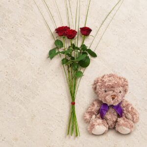 Tre röda rosor med en gammelrosa nalle med lila rosett